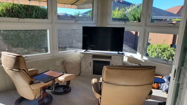 Een zonnige kamer met een televisie, twee leren fauteuils, een tafeltje en grote ramen met uitzicht op een tuin.