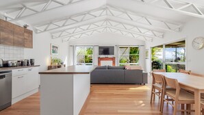 A modern kitchen with a central island, a dining area with a wooden table and chairs, a grey sofa, and a fireplace. - Bush to Beach Family & Fur Coastal Retreat (Moruya Heads)