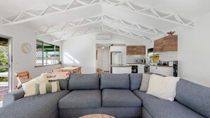 A modern living room with a grey sectional sofa, a dining area with a wooden table, and a kitchen with white cabinets and a wooden accent wall. - Bush to Beach Family & Fur Coastal Retreat (Moruya Heads)