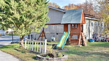 Un terrain de jeux avec un toboggan et des balançoires, un arbre et un bâtiment dont le mur est en briques, visible au fond.