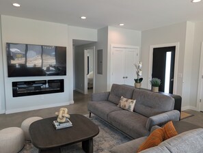 A modern living room with a flat-screen TV, a gray sofa, a coffee table, and a potted plant. - New build Luxe escape w Heated Pool, Hot Tub, Fire Pit, State Park and Beaches (Stevensville)
