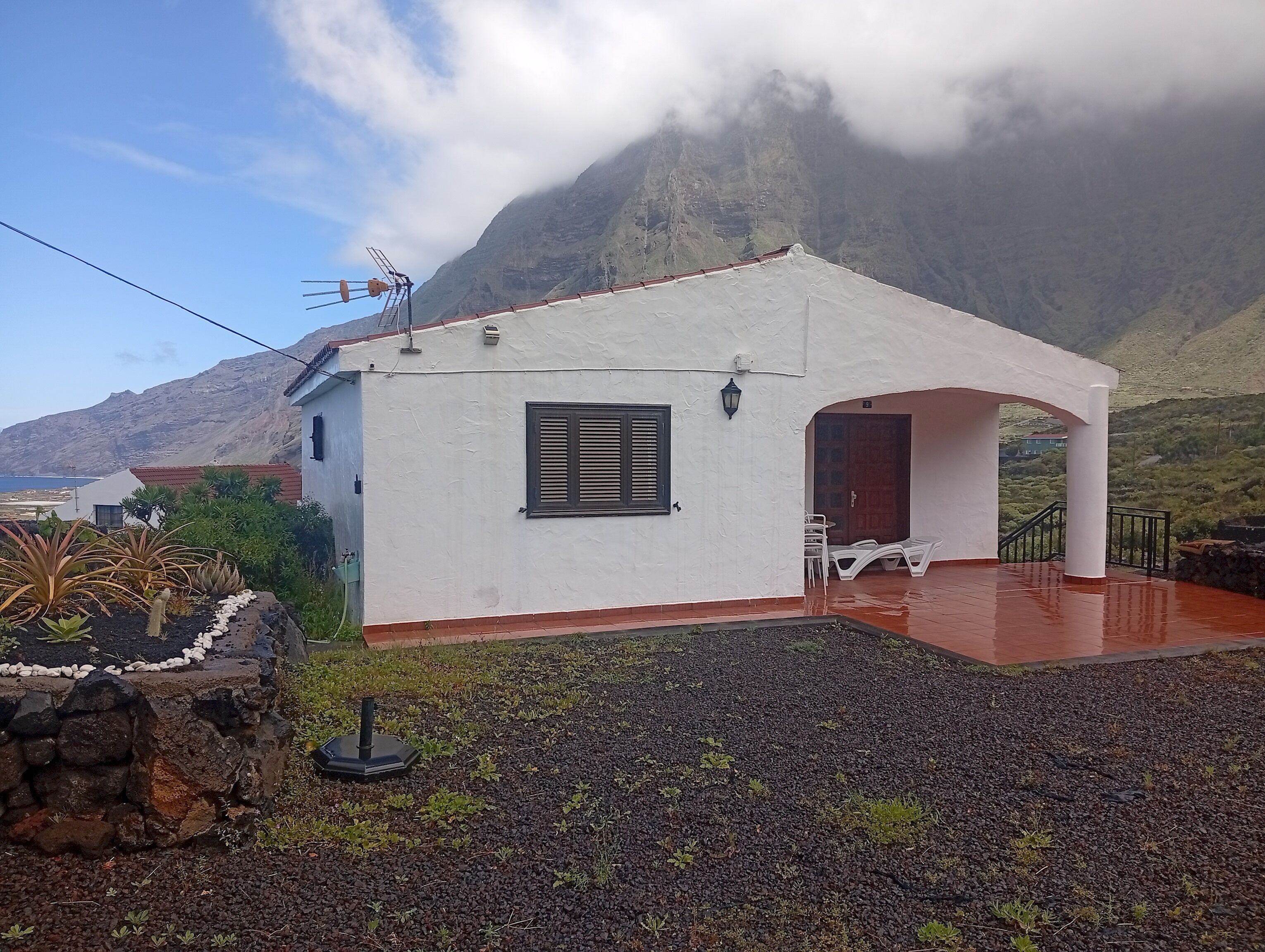 A white building with a small porch, a window with shutters, and a stone wall with plants.