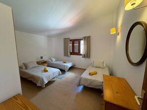 Une chambre avec trois lits simples, une table de chevet en bois, une petite table, un miroir et une fenêtre avec des rideaux.