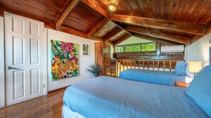 Een kamer met een houten plafond, een bed met een blauwe sprei, een schilderij aan de muur en een raam met uitzicht op bomen.