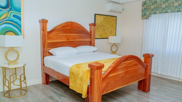 A wooden bed with a headboard, two bedside tables with lamps, and a window with curtains. - Royale Jazz Hotel  (Kingston)