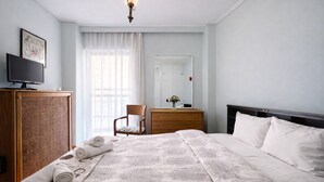 Ett hotellrum med en säng, en byrå i trä, en stol, ett skrivbord, en tv och ett stort fönster med gardiner.