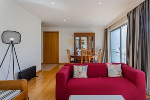 A living room with a red sofa, a dining table with chairs, a wooden door, and a large window with curtains.