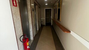A hallway with a fire extinguisher, a fire alarm, and a door at the end. - Flat em Moema com Cozinha (São Paulo)