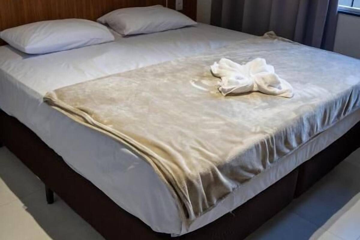 Een net opgemaakt bed met een witte dekbedovertrek en een beige deken, een houten hoofdbord en een telefoon naast het bed.