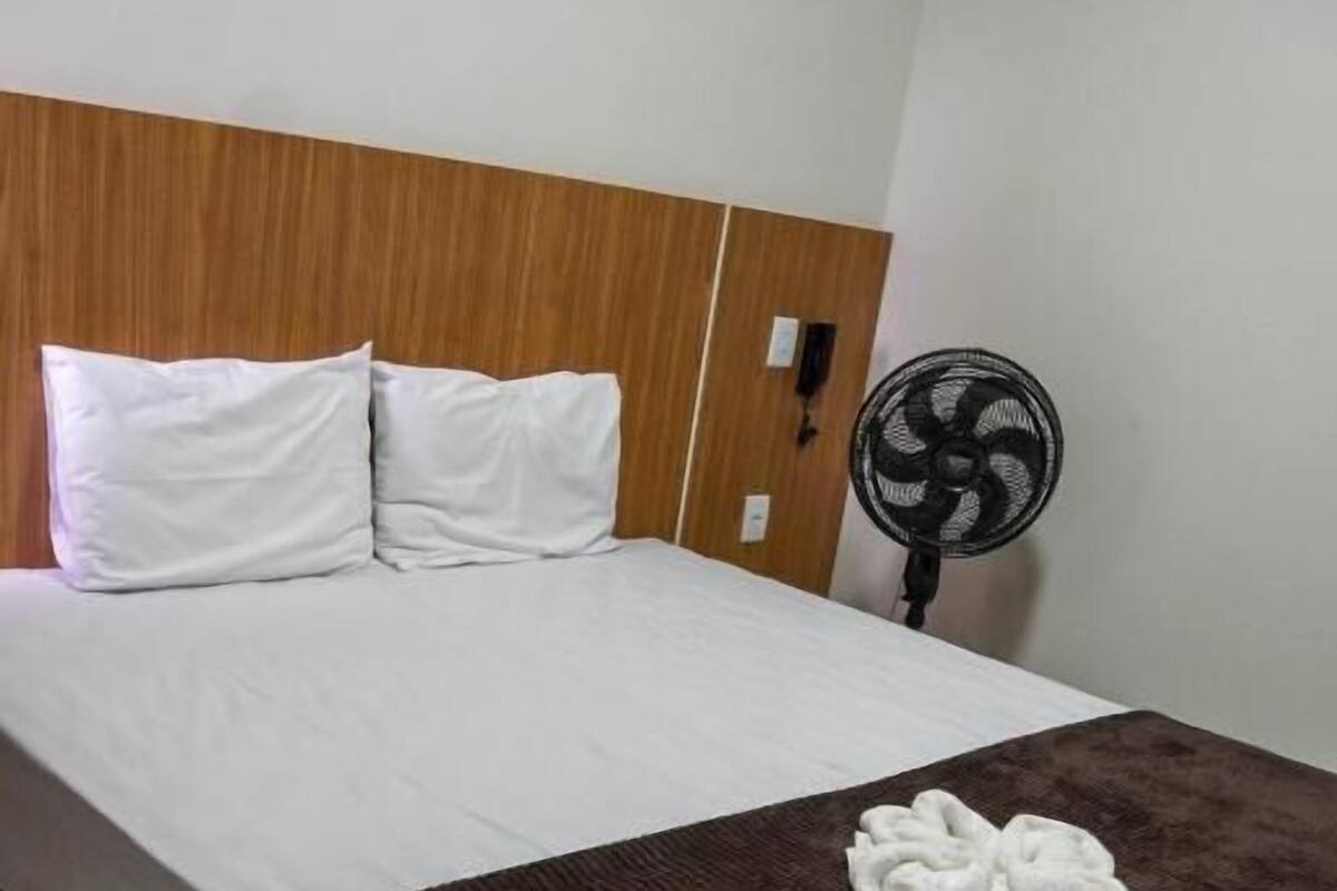 Een hotelkamer met een bed, witte kussens, een bruine deken en een ventilator.