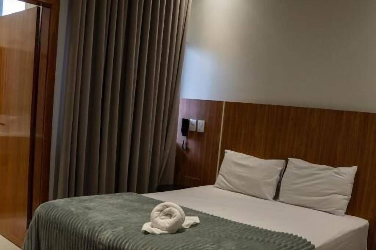 Een hotelkamer met een bed, gordijnen, een airconditioner en een houten hoofdeinde.