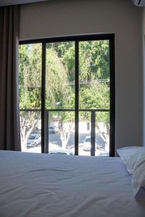A hotel room with a large window, a bed, and a view of trees and parked cars outside. - Norte Hotel (Taiobeiras)