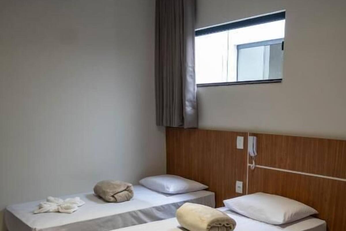 Een hotelkamer met drie eenpersoonsbedden, elk met een handdoek en kussen, een houten hoofdbord, een raam met gordijnen en een airconditioner aan de muur.