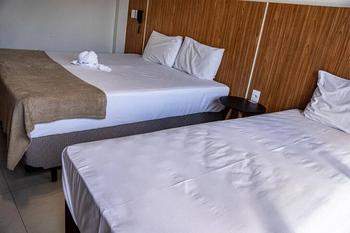 Twee bedden met witte lakens, een bruine deken op een ervan, en een klein houten nachtkastje met een telefoon.