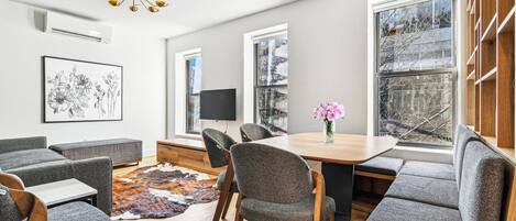 Un salon moderne avec une table à manger en bois, des chaises à revêtement gris, une grande fenêtre offrant une belle vue et un téléviseur à écran plat fixé au mur.