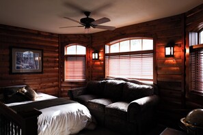 A log cabin room with a bed, a sofa, a ceiling fan, and two windows with blinds. - Lakefront Home • Sleeps 19+ • Perfect for Groups & Families (Turtle Lake)