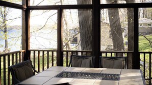Un espacio al aire libre cubierto con una mesa y sillas, ventanas amplias y vistas a árboles y una casa.