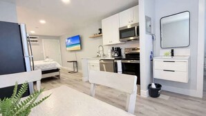 Een moderne keuken met witte kastjes, een magnetron en een kookplaat. Er staat een eettafel met stoelen, een televisie hangt aan de muur, en er is een bed op de achtergrond.