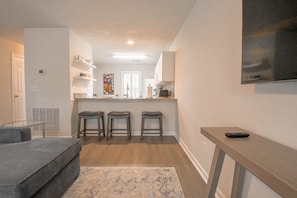 Une cuisine moderne avec un îlot central, des tabourets de bar, un canapé gris et un téléviseur fixé au mur.