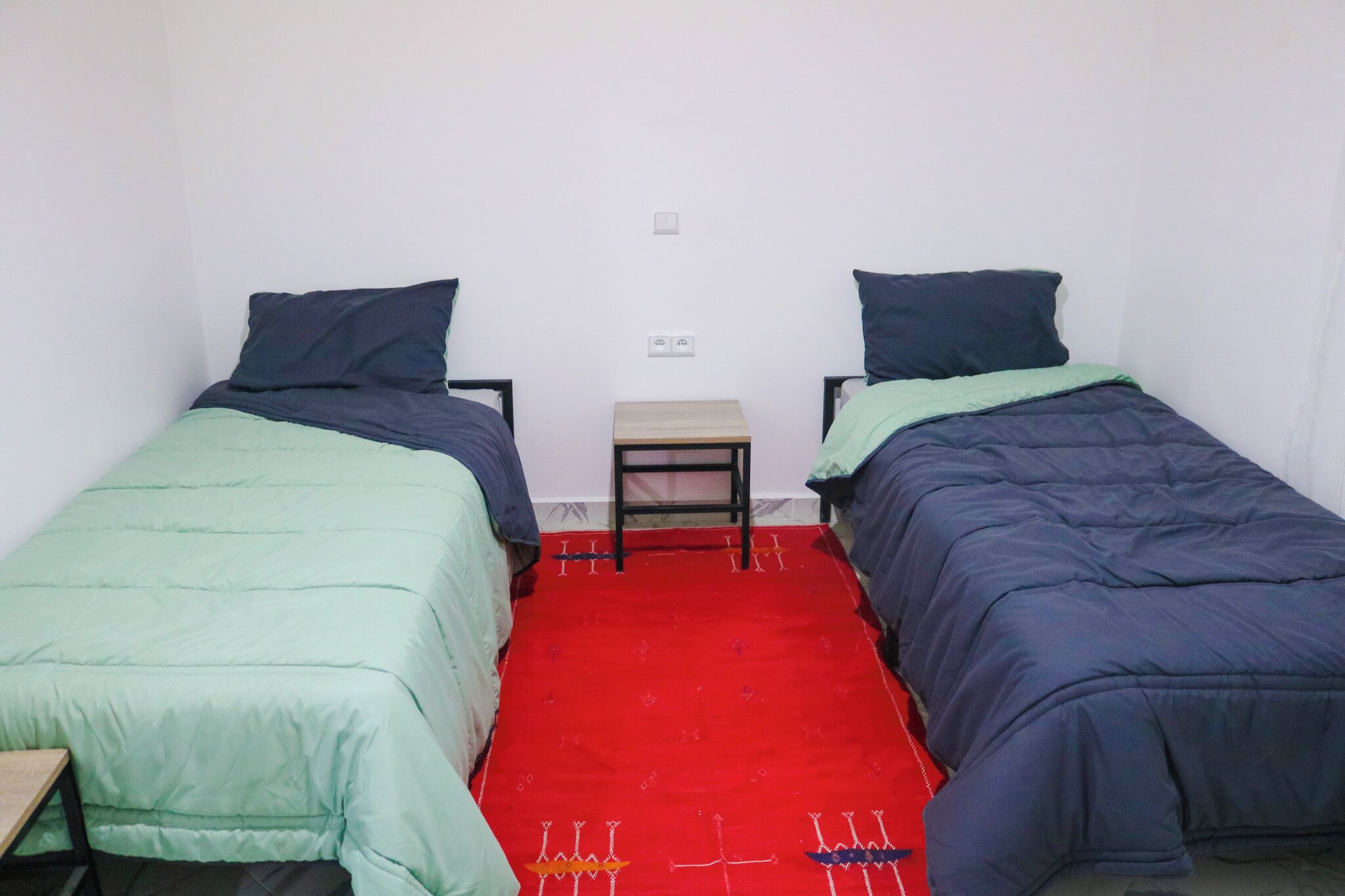 Une petite chambre avec deux lits simples, une petite table de chevet en bois et un tapis rouge.