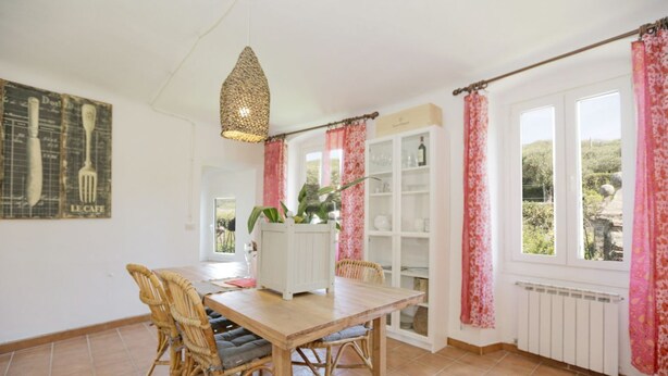Uma área de refeições com mesa de madeira, cadeiras de vime, luminária pendente, armário branco e janela com cortinas cor-de-rosa.