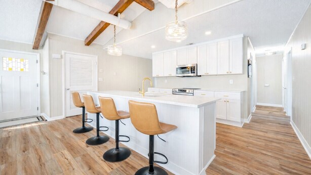 A modern kitchen with white cabinetry, a central island with bar stools, and wooden flooring. - New! Beach Cottage, Pool, PickleBall, Swim, Surf (Virginia Beach)