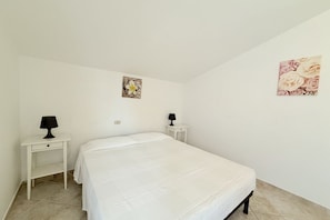 A single-bed room with a white bedspread, two bedside tables with lamps, a wall-mounted artwork, and a tiled floor.
