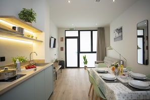 Eine moderne Küche mit Spüle, Herd und Arbeitsplatte. Es gibt einen Essbereich mit einem Tisch für zwei Personen, einer Couch und einem großen Fenster mit Vorhängen.