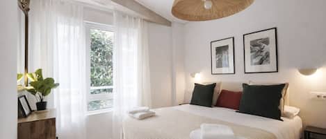 Un dormitorio con una cama grande, una lámpara colgante, un ventanal con cortinas translúcidas y cuadros en la pared.
