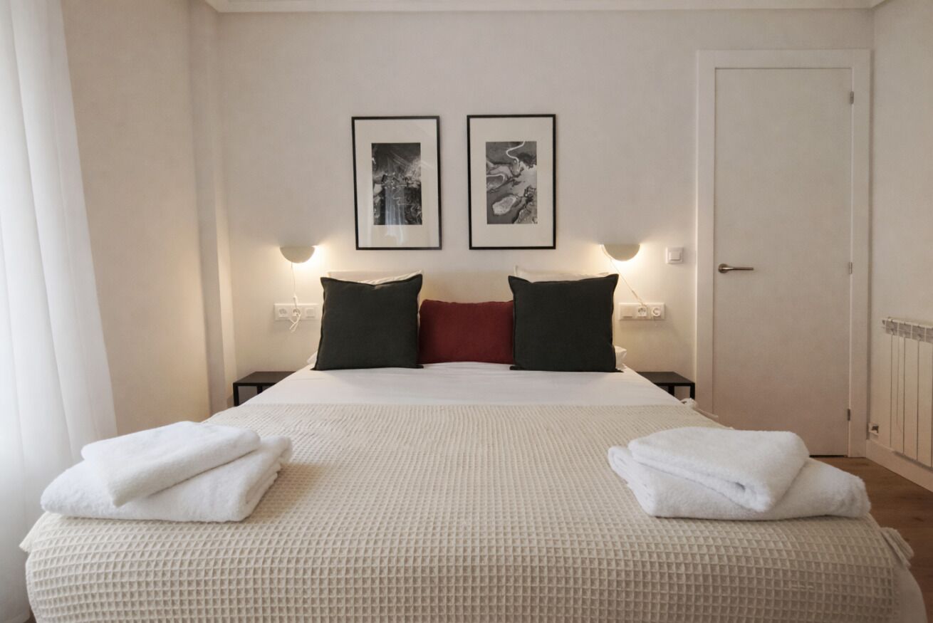 Una habitación de hotel con una cama, dos cuadros en la pared, una puerta y una mesita de noche con una lámpara.