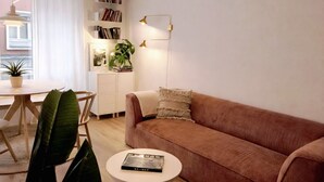 Ett vardagsrum med en soffa, ett soffbord med en bok på, en bokhylla, en växt och en lampa.