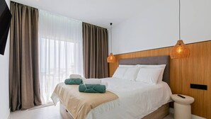 Ein modernes Hotelzimmer mit einem großen Bett, zwei Nachttischlampen, einem Nachttisch und Blick auf die Stadt durch große Fenster.