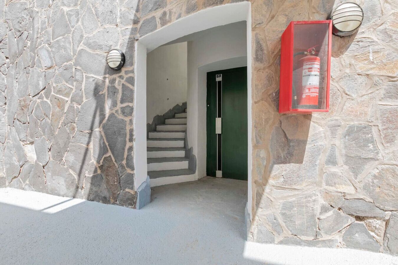 Een stenen buitenmuur, een trap die naar een deur leidt, en een rode brandblusser aan de muur bevestigd.
