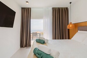 Quarto de hotel com uma cama, duas toalhas enroladas, uma mesinha de cabeceira, uma cadeira, uma TV na parede e vista para o mar pela janela.