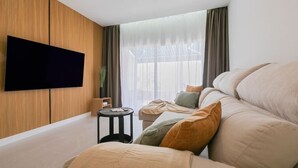 Ein modernes Wohnzimmer mit einem Wand-Fernseher, einer Couch mit Kissen, einem kleinen runden Tisch und einem großen Fenster mit Vorhängen.