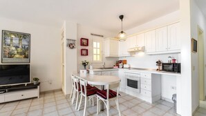Une cuisine compacte avec des meubles blancs, une table à manger avec des chaises, un téléviseur et un luminaire suspendu.