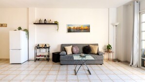 Un salon comprenant un canapé, une table basse, un réfrigérateur blanc, une étagère avec des objets décoratifs et un tableau accroché au mur.