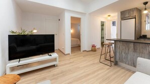 Un apartamento moderno con una televisión de pantalla plana sobre un soporte blanco, una zona de cocina con encimera de madera y taburetes de bar, y un dormitorio visible a través de una puerta abierta.