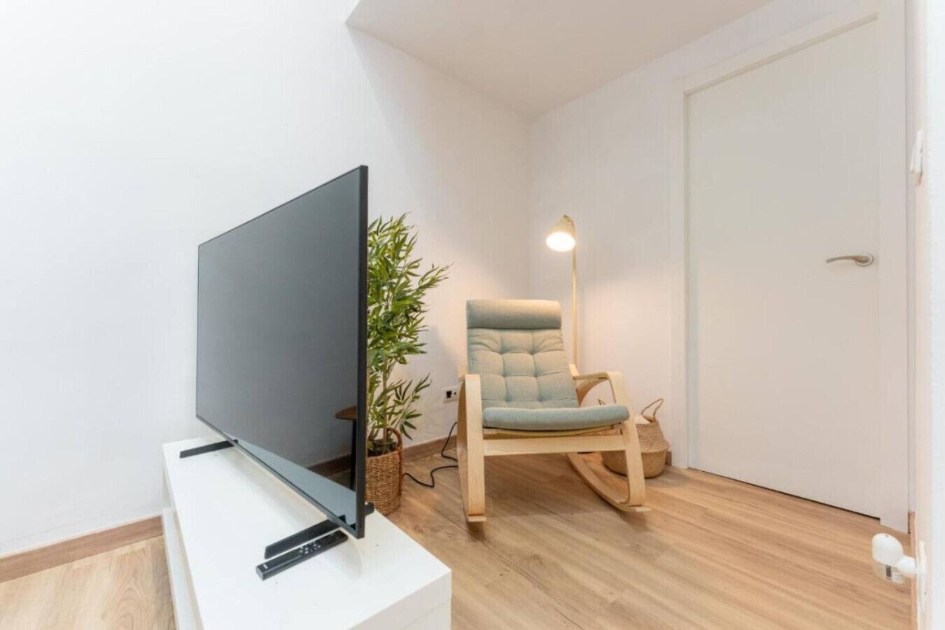 Een kamer met een flatscreen televisie op een witte standaard, een houten vloer, een lichtkleurige stoel, een potplant en een witte deur.