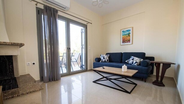 A living room with a sofa, a coffee table, a fireplace, and a sliding glass door leading to an outdoor area. - Electra (Aigialeia)