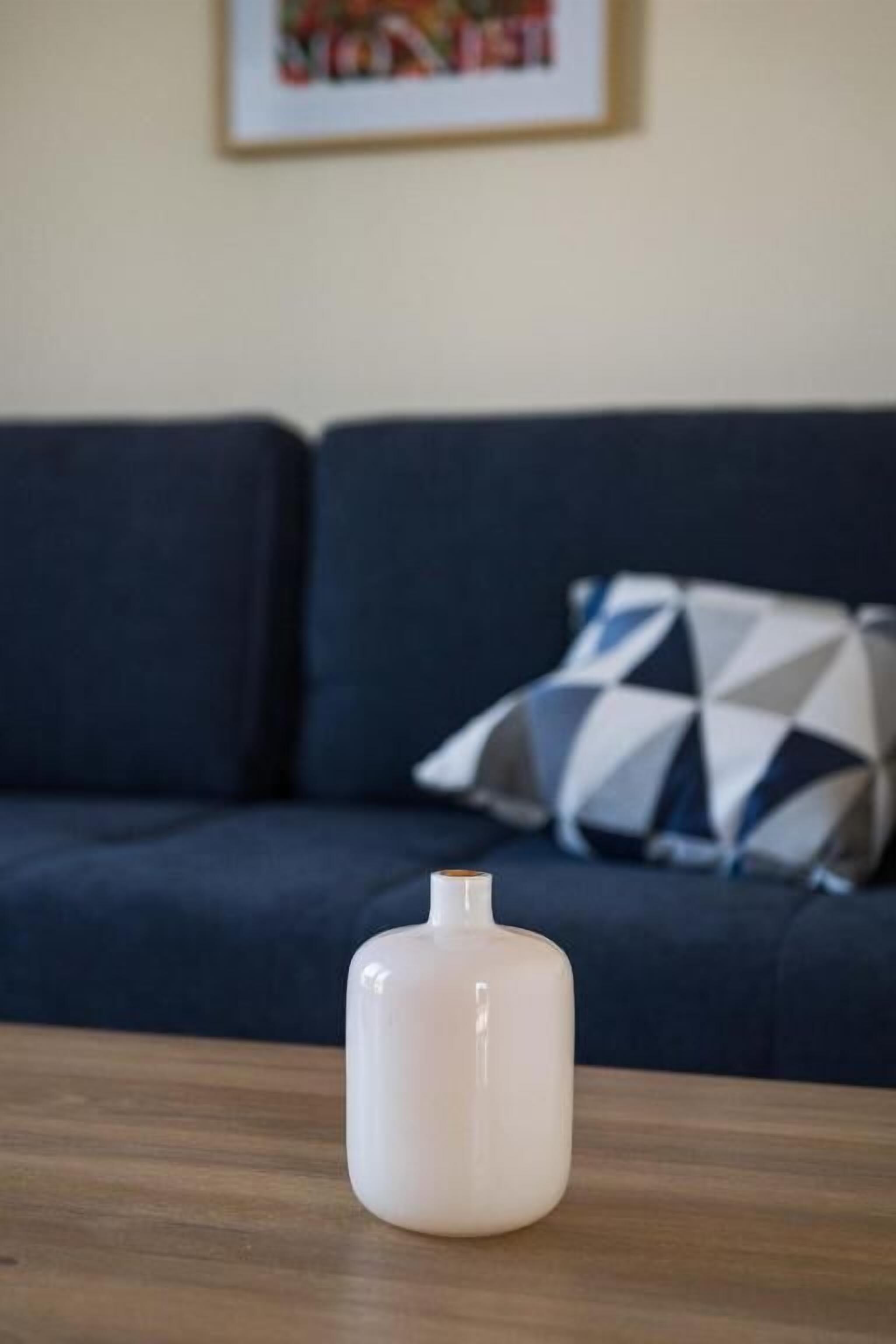 Een witte vaas op een houten tafel, met een donkerblauwe bank en gekleurde kussens op de achtergrond.