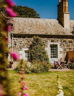 A stone house with a green door, a chimney, and a garden with flowers and a table. - The Redwing - Two Bedroom Villa, Sleeps 4 (Portpatrick)