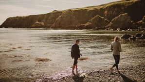 Deux personnes se promènent sur une plage rocheuse, avec une falaise en arrière-plan.