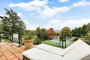 Un balcón con vistas a una cancha de tenis, una planta grande en maceta y una zona de estar con cojines blancos.