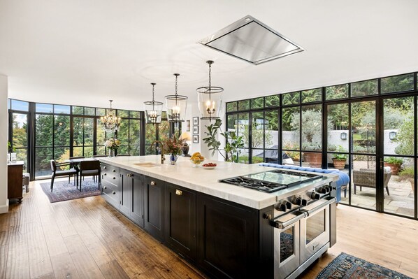 A modern kitchen with a large island, stainless steel appliances, and large windows offering a view of the outdoors.