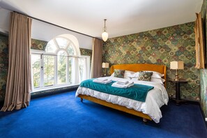A bedroom with a large bed, a window with curtains, patterned wallpaper, and a chandelier.