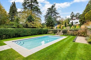 A rectangular swimming pool surrounded by well-maintained grass and hedges, with a house in the background.