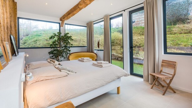 A bedroom with a large bed, a chair, a plant, and a view of the outdoors.