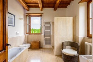 Une salle de bain avec une baignoire, un lavabo, une chaise en osier, une fenêtre donnant sur un paysage agréable et un porte-serviettes.