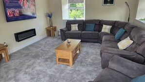 A living room with a sectional sofa, a flat-screen TV, a wooden coffee table, and a window with a view of greenery. - Farm Cottage at Foot Angus Glens (Kirriemuir)